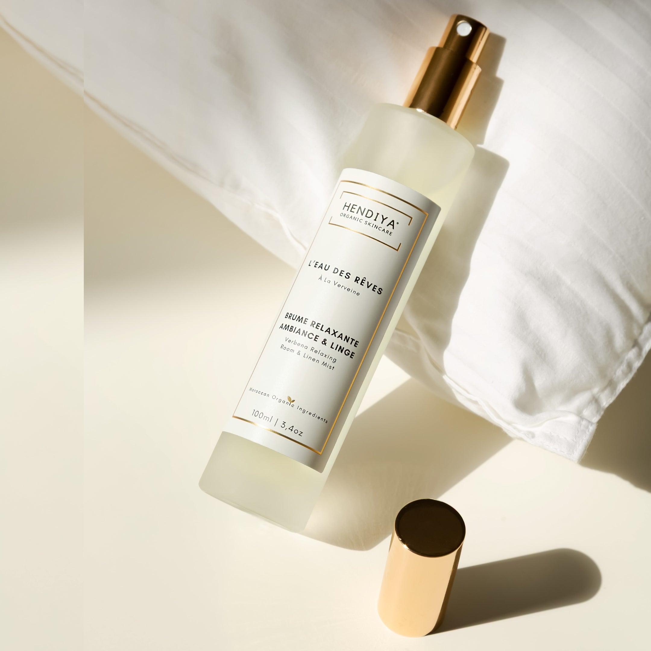 Flacon de la brume relaxante L'Eau des Rêves Hendiya à la verveine citronnée, posé sur un oreiller et du linge de lit, pour une utilisation en ambiance et sur le linge.