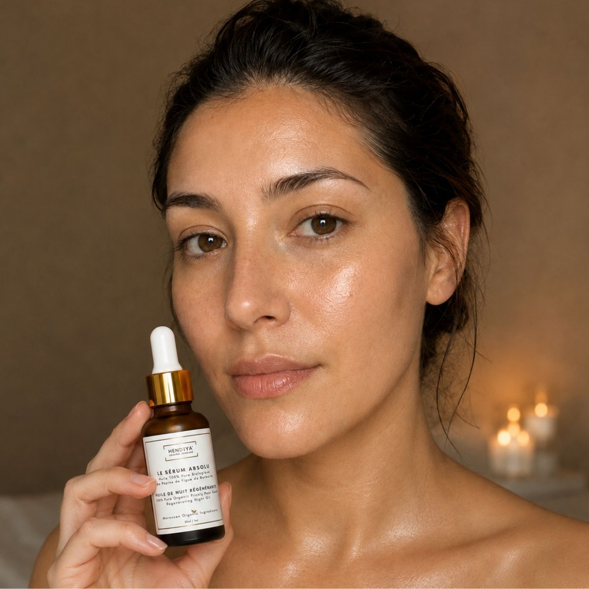 Femme tenant le flacon Le Sérum Absolu Hendiya dans une ambiance de rituel du soir aux chandelles, huile de pépins de figue de barbarie